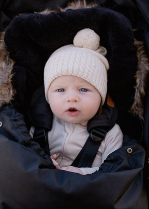 Baby in a black footmuff and white knitted hat with pom-pom.