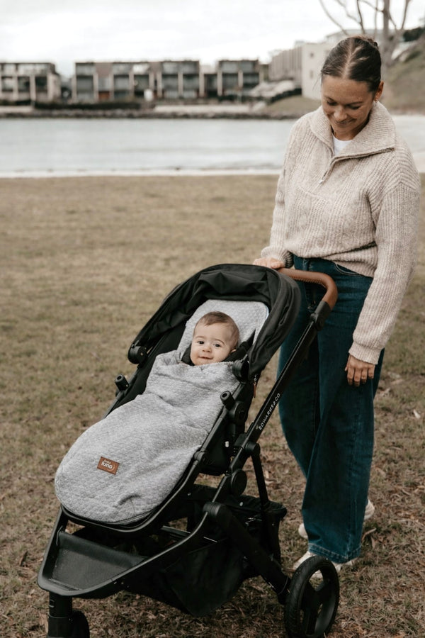Woman pushing a stroller with a baby in a gray summer footmuff by the ocean