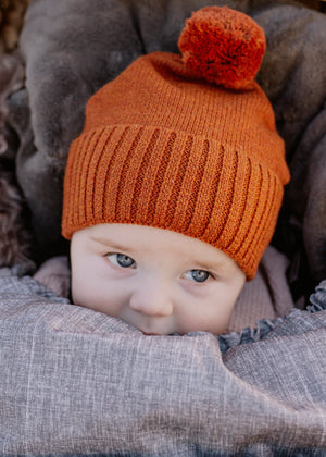 Baby wearing a rust brown knit hat with a pom-pom, peeking out from behind Chic Grey baby luno footmuff