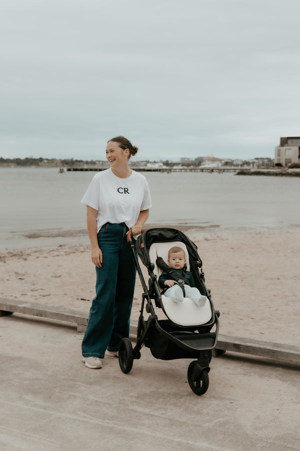 Mother pushing a stroller with summer lite footmuff pram liner with a child on a boardwalk by a waterfront.