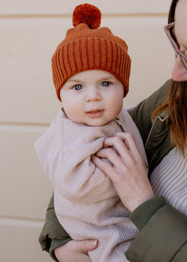 Baby wearing a brown rust knit hat with a pom-pom, held by mum.