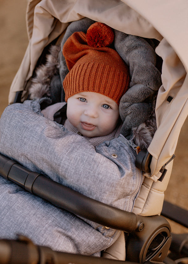 Baby in baby luno footmuff in a stroller wearing rust knit hat with a pom-pom.