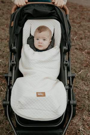 Baby in a stroller wrapped in a cream baby luno Lite Summer Footmuff blanket outdoors.