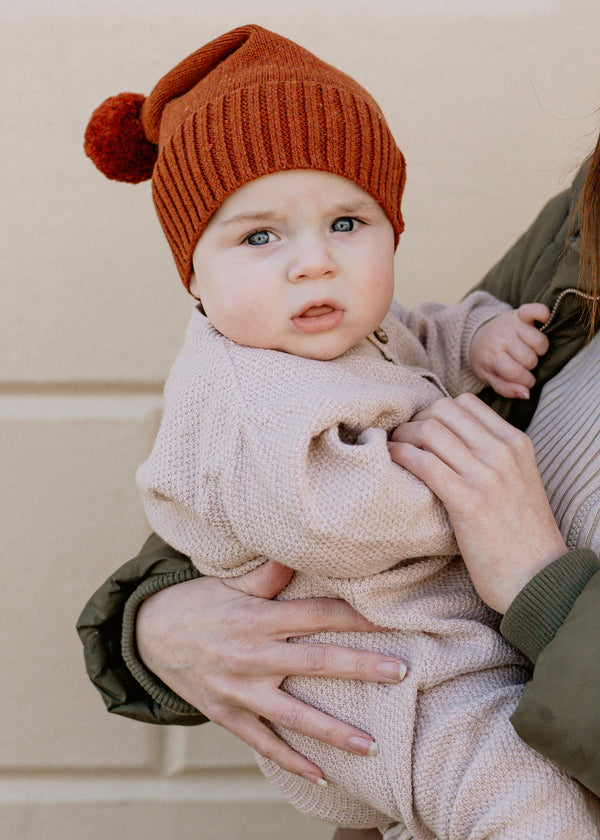 Baby wearing baby luno brown knit hat and beige sweater being held by mum.
