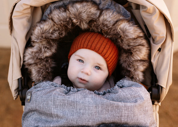 Baby in Chic Grey baby luno footmuff wearing rust orange knit hat