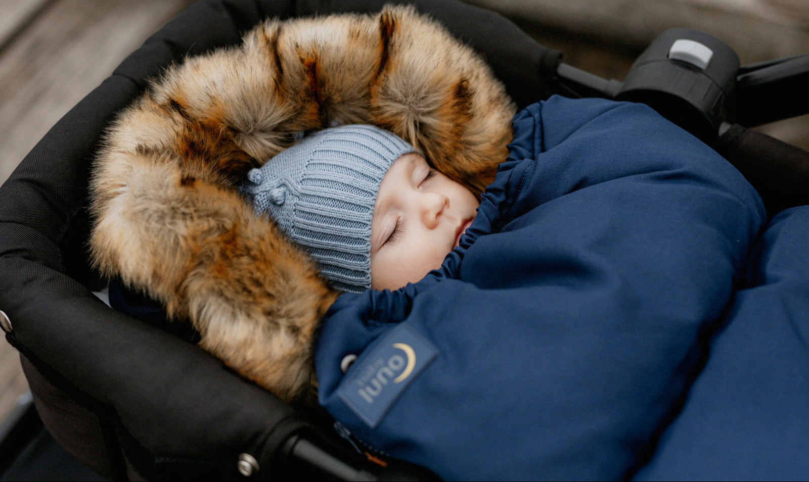 A baby in a deep blue pram liner with faux fur trim, wearing a blue beanie with a detachable bow.