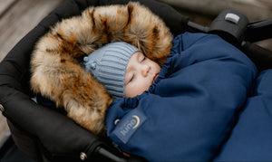 A baby in a deep blue pram liner with faux fur trim, wearing a blue beanie with a detachable bow.