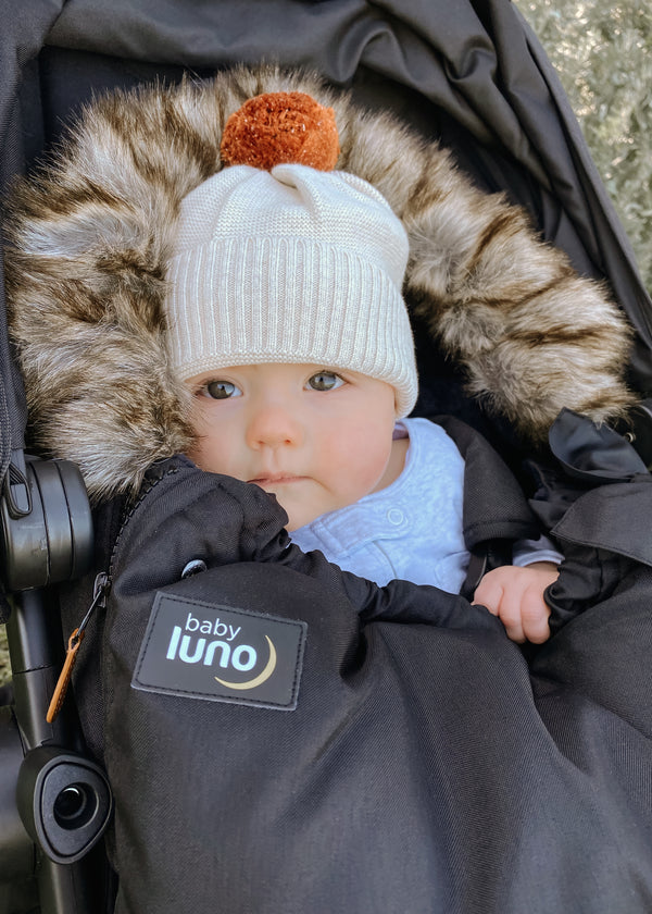 A baby in a black footmuff pram liner with gray/brown fur, wearing a white beanie with a pom-pom, in a stroller.